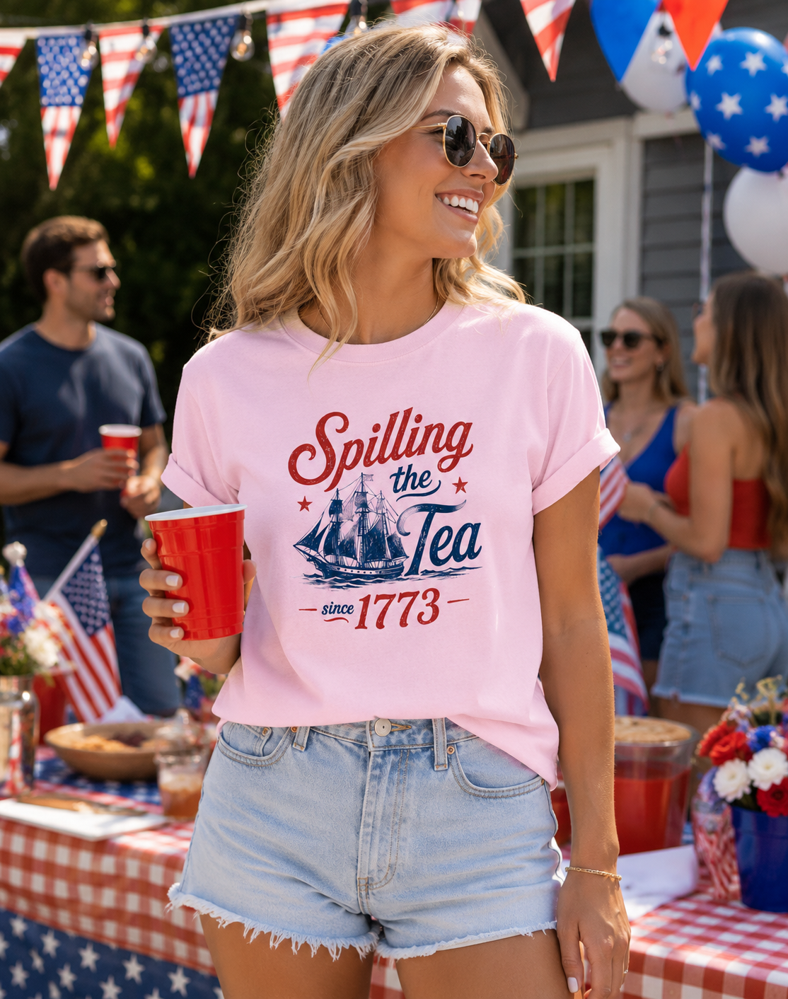 smiling woman wearing light pink patriotic graphic tee with spilling the tea since 1773 quote and vintage colonial ship design at festive fourth of july barbecue party