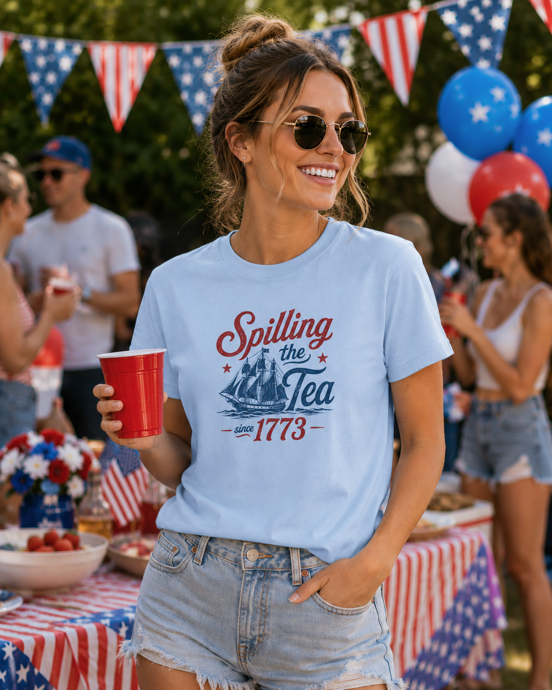 stylish woman wearing light blue patriotic graphic tee with spilling the tea since 1773 quote and vintage ship design at festive fourth of july backyard barbecue party